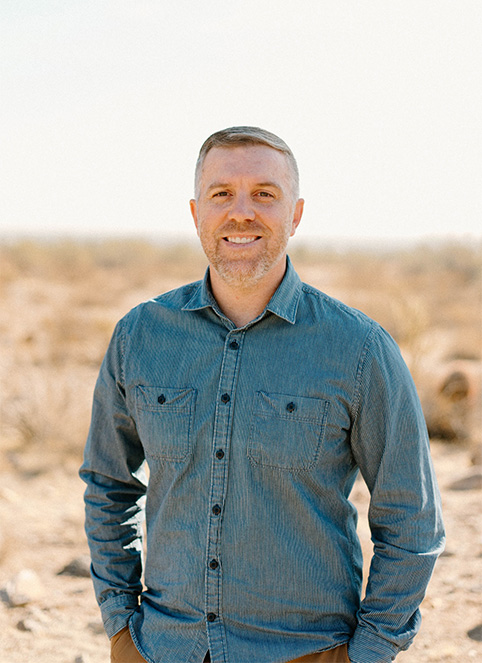 Portrait of Dr. Caleb Goodman in a denim shirt, standing outdoors in a dry landscape.